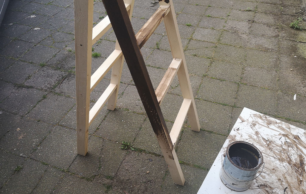 A wooden ladder is partially painted with dark wood stain, an open paint can is next to it.
