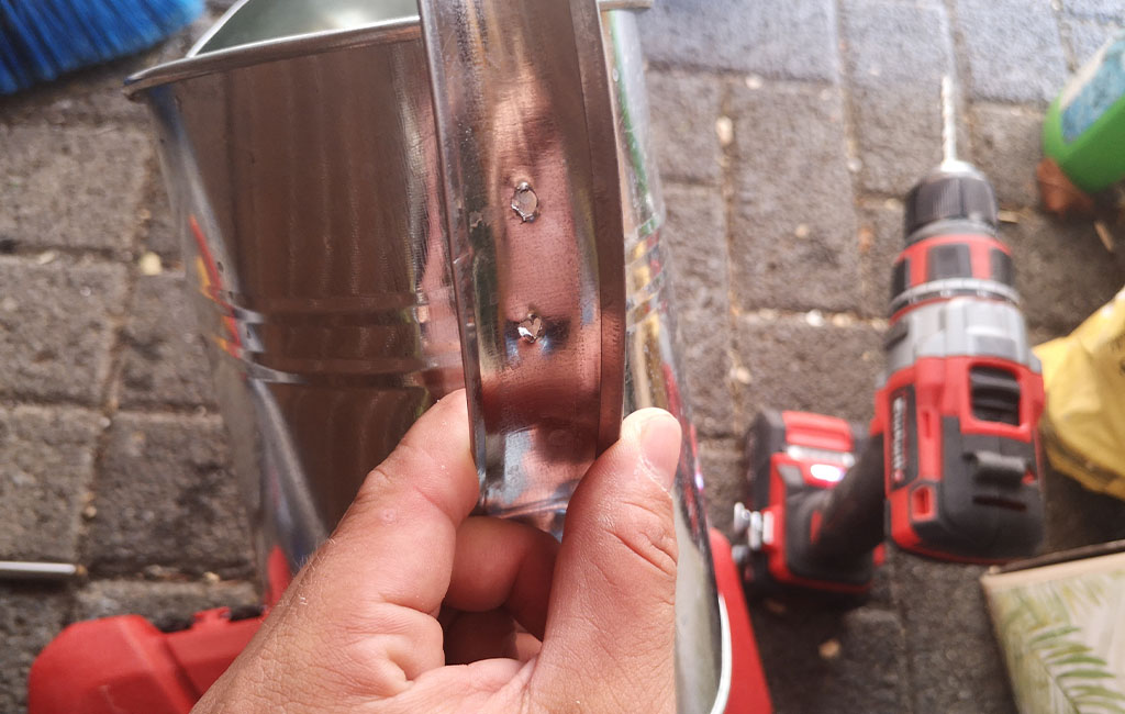 A hand holds a metal watering can with drilled holes, an Einhell cordless drill is in the background.