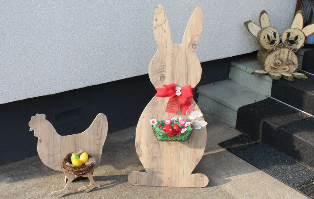 Decorative wooden figures of a rooster and a bunny with Easter eggs in front of a house wall.