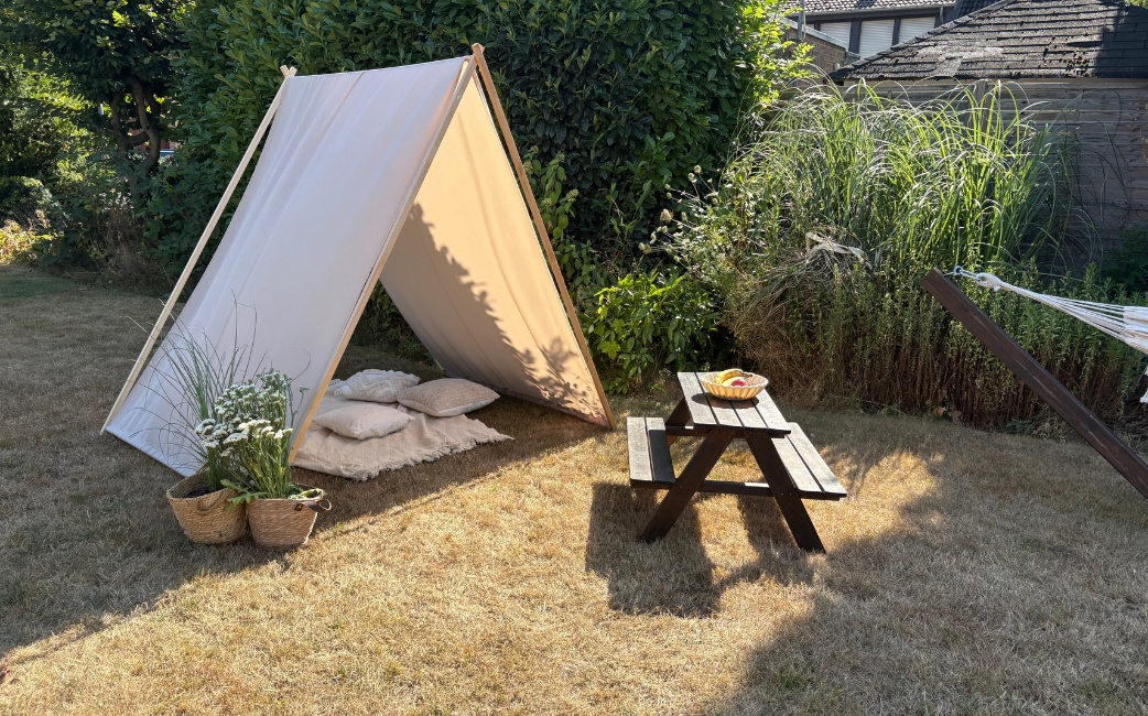 Beige fabric stretched over an A-shaped wooden frame as a sun shelter in a garden, with a picnic table and plants nearby.