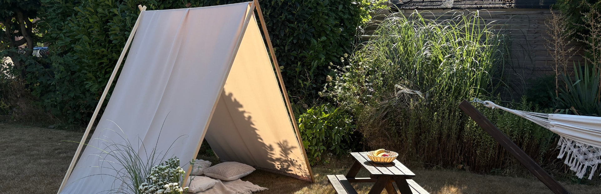 Beige fabric stretched over an A-shaped wooden frame as a sun shelter in a garden, with a picnic table and plants nearby.