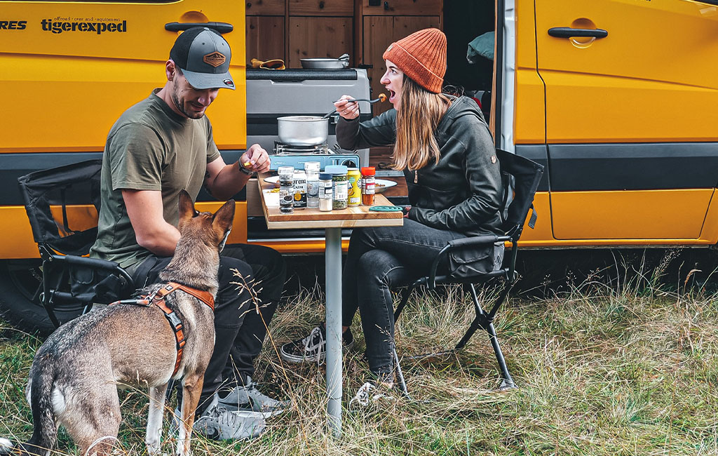 A man and woman eat at a camping table in front of a yellow van, with a dog standing nearby.