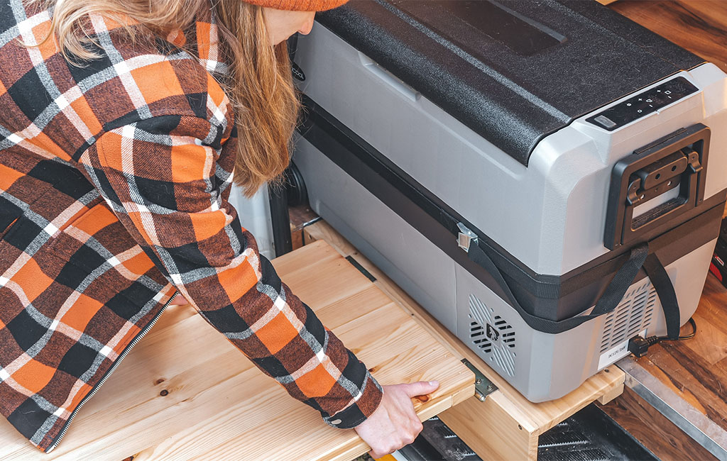 A woman pulls out a foldable wooden board next to a large cooler inside the camper.
