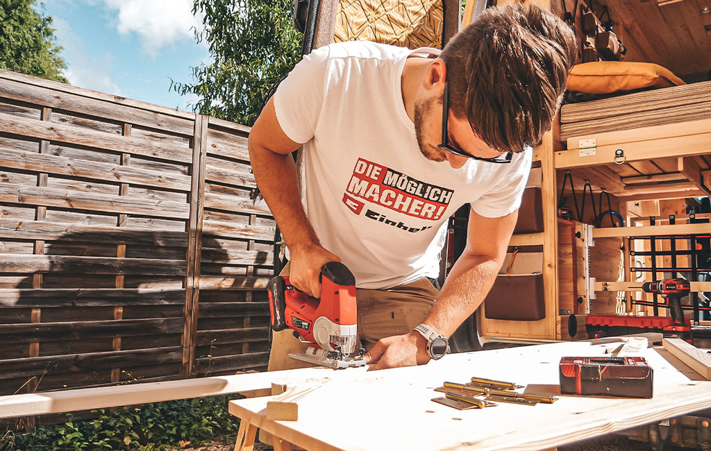 A man wearing an Einhell T-shirt uses a jigsaw to cut a wooden board in front of a camper.
