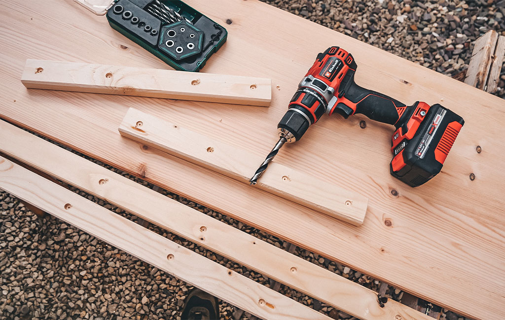 A wooden board with pre-drilled strips, next to a cordless drill and a drill bit set.