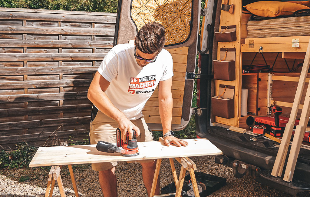A man sands a wooden board with an Einhell sander on sawhorses in front of the camper.
