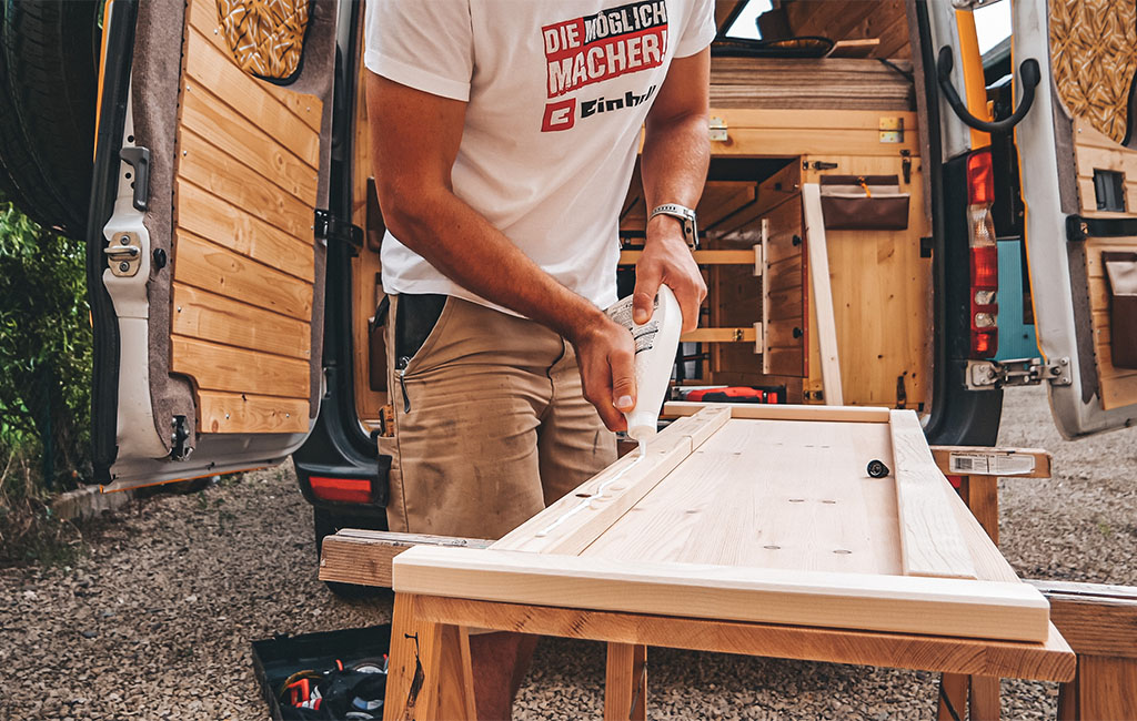 A man applies wood glue to a wooden board resting on sawhorses in front of the camper.