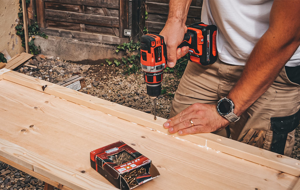 A man screws a wooden strip onto a glued wooden board using a cordless screwdriver.