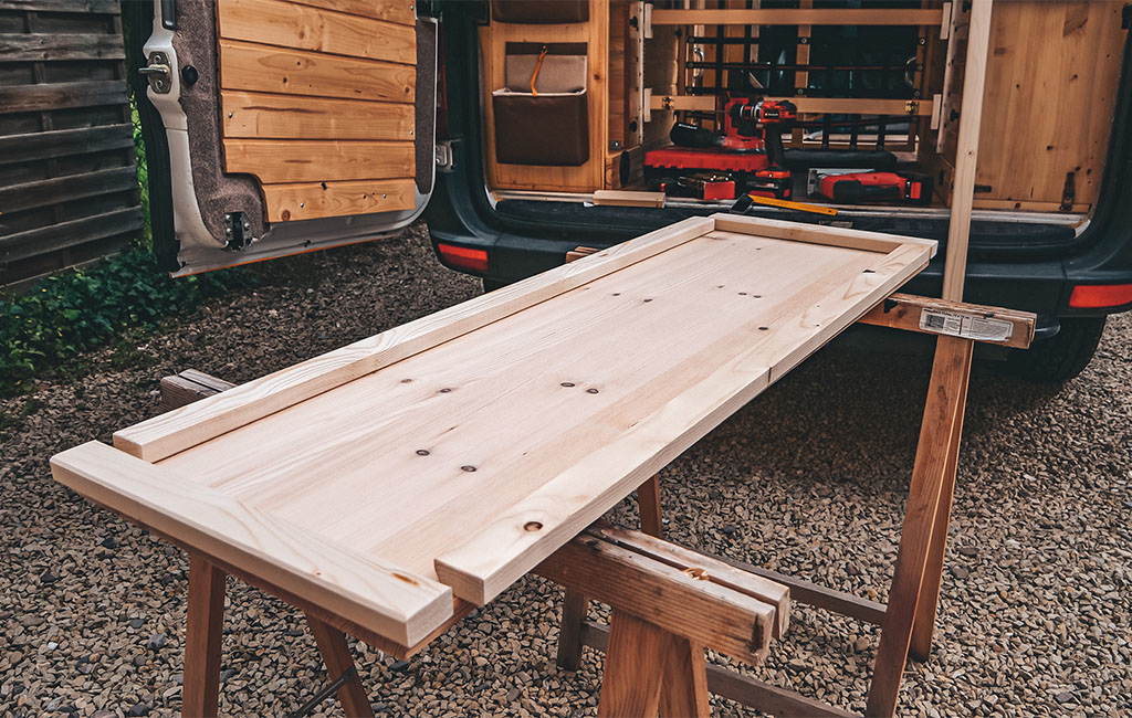 A large, light-colored wooden board lies on two wooden sawhorses in front of an open camper with tools.