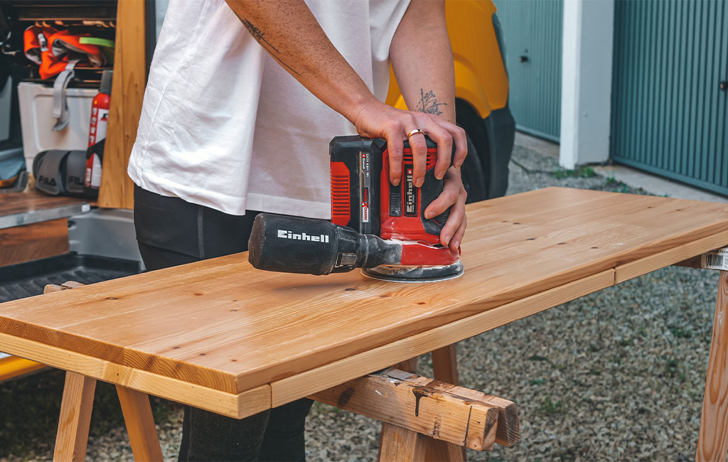 A person sands a wooden board with an Einhell orbital sander placed on two wooden sawhorses.