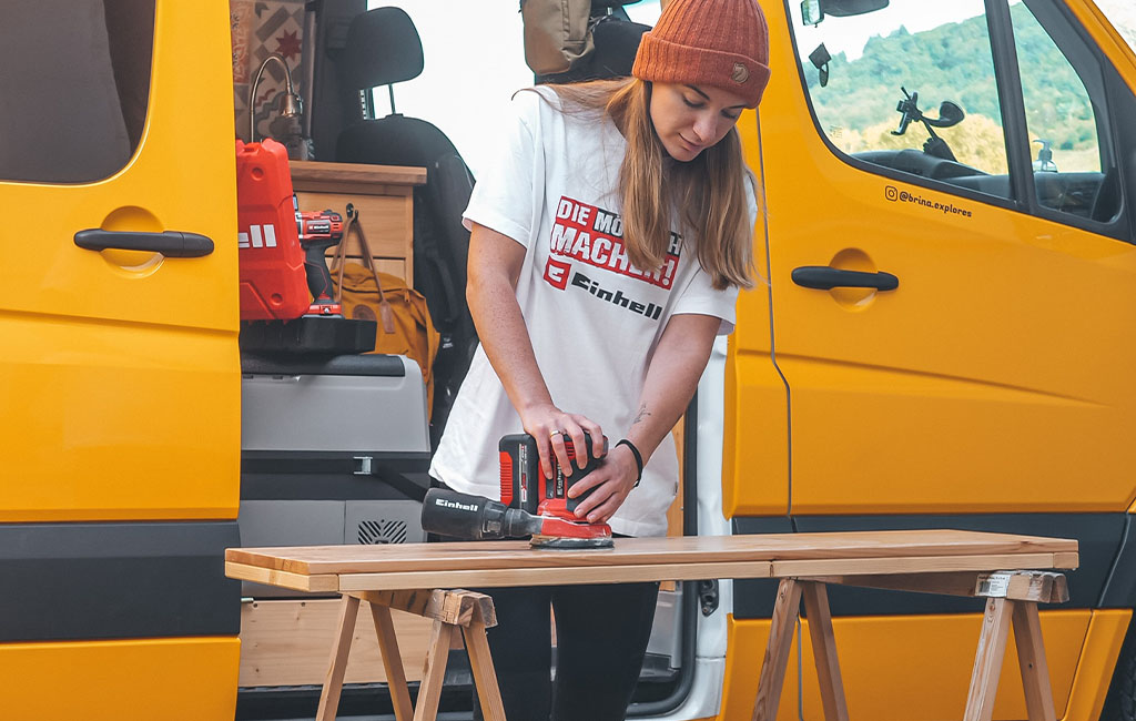 A woman sands a wooden board outside a yellow camper using an Einhell sander.