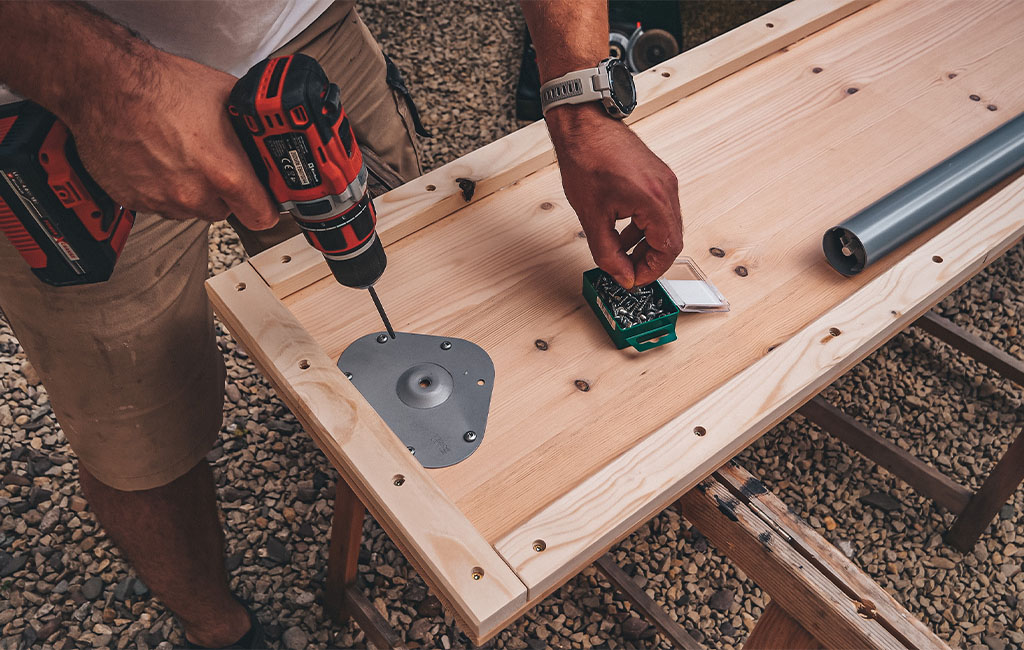 A man uses a cordless drill to screw a round table leg bracket underneath a wooden board.