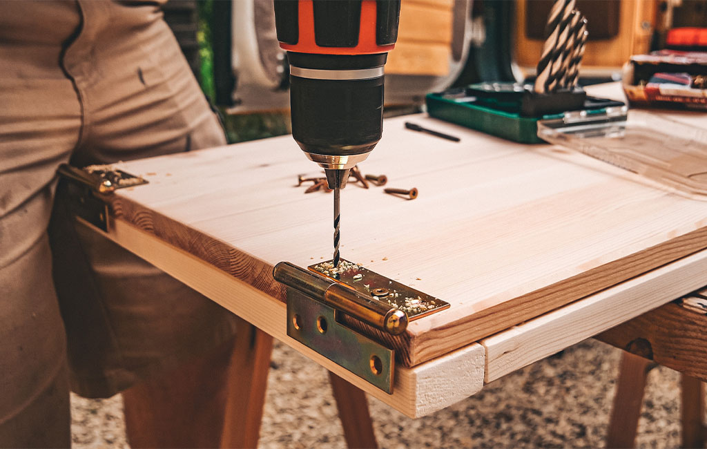 A metal hinge is being attached to the corner of a wooden board using a cordless drill.