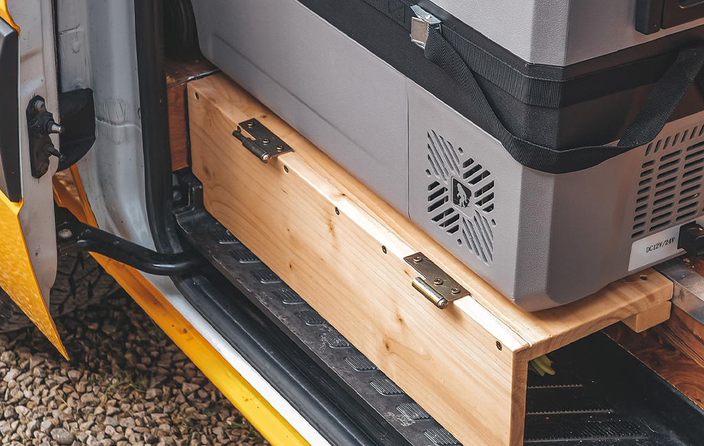 A folded wooden board with hinges is stored next to a cooler inside the camper.