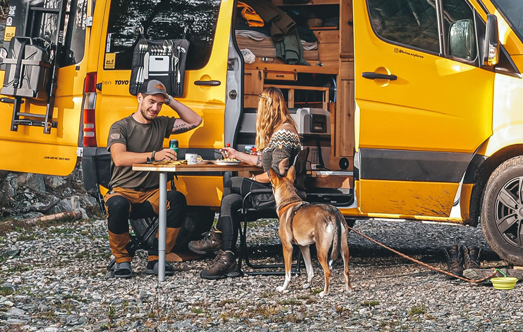 A couple sits at a folding table in front of an open camper, with a dog standing nearby.