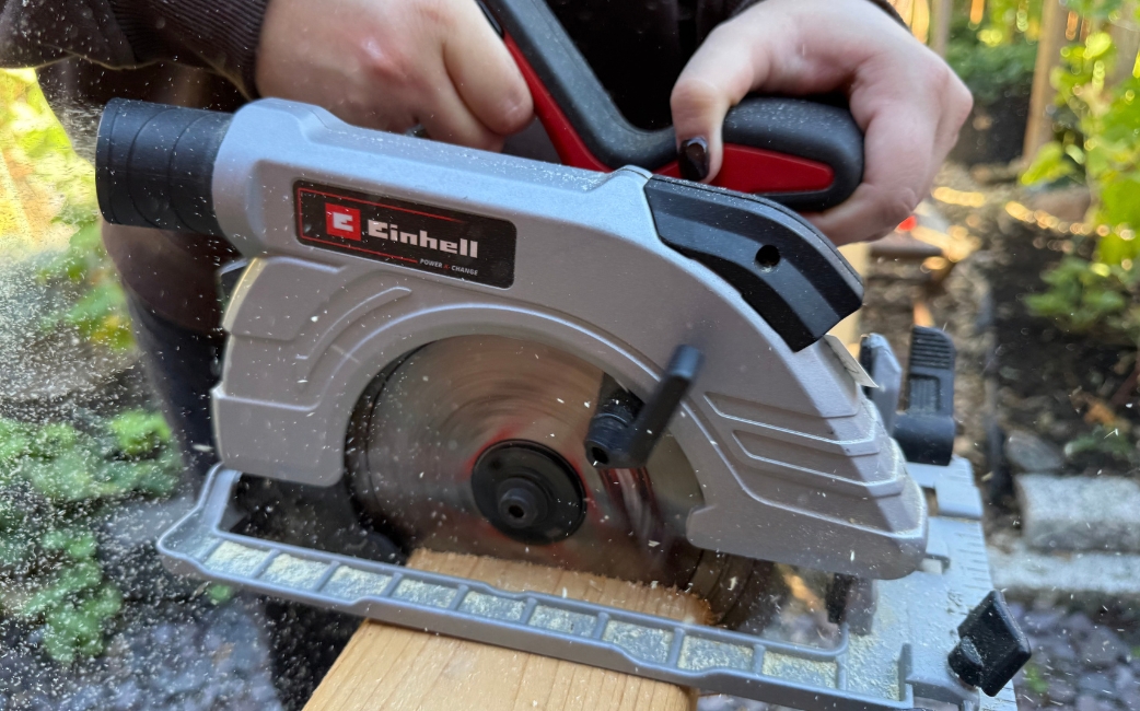 Nahaufnahme einer Einhell-Kreissäge, die ein Holzbrett zersägt, wobei Sägespäne herumfliegen.