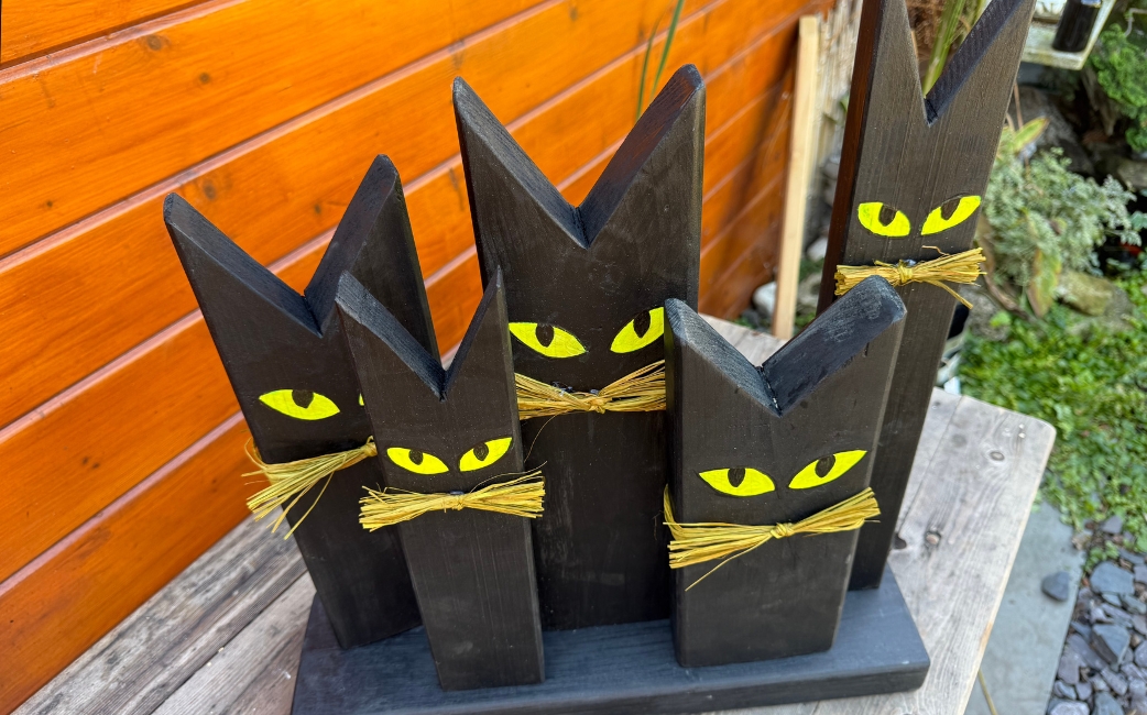 Fünf schwarze, katzenförmige Holzfiguren mit gelben Augen und Schnurrhaaren aus Stroh stehen auf einem gemeinsamen Holzsockel.