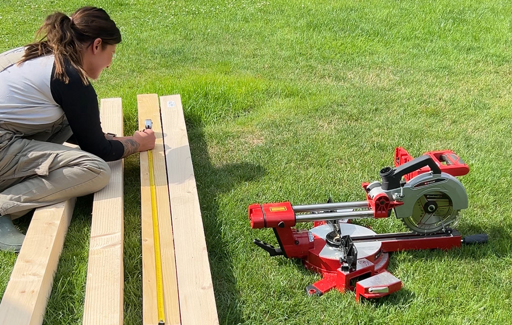 A woman kneels on the grass and measures wooden beams with a tape measure; next to her is an Einhell mitre saw.