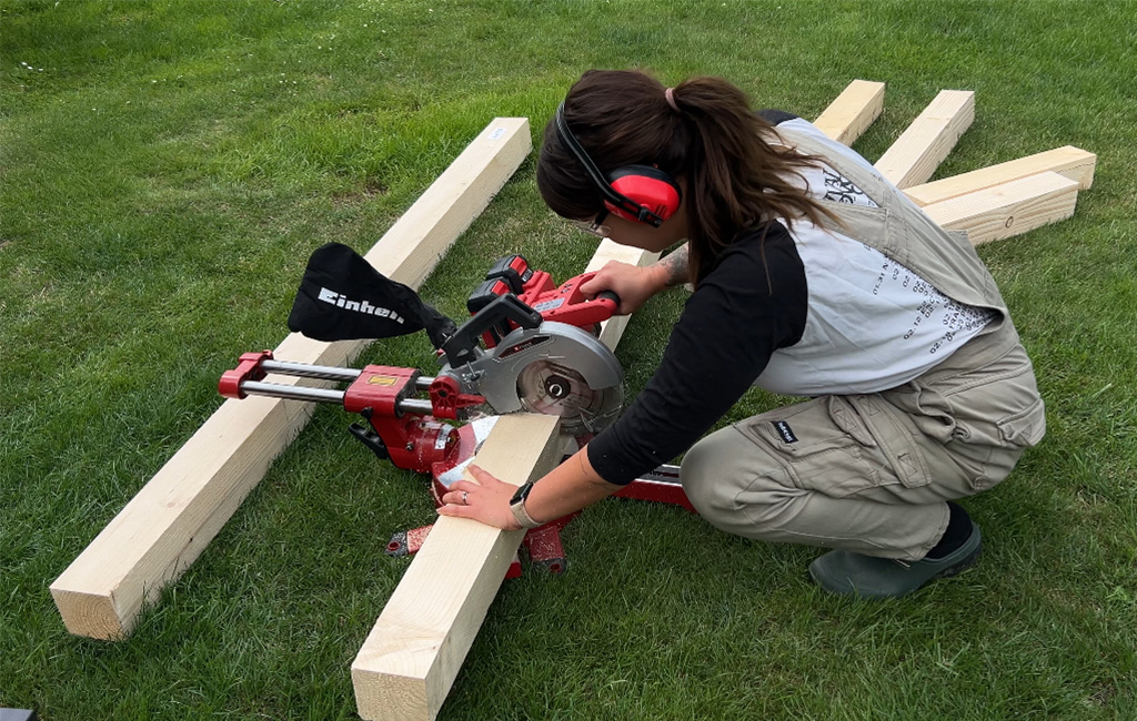 A woman cuts a squared timber with an Einhell mitre saw on a lawn; she is wearing hearing protection.