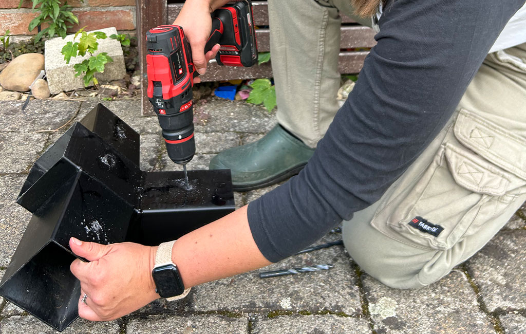 A woman drills holes in a black metal bracket using an Einhell cordless drill on a paved surface.
