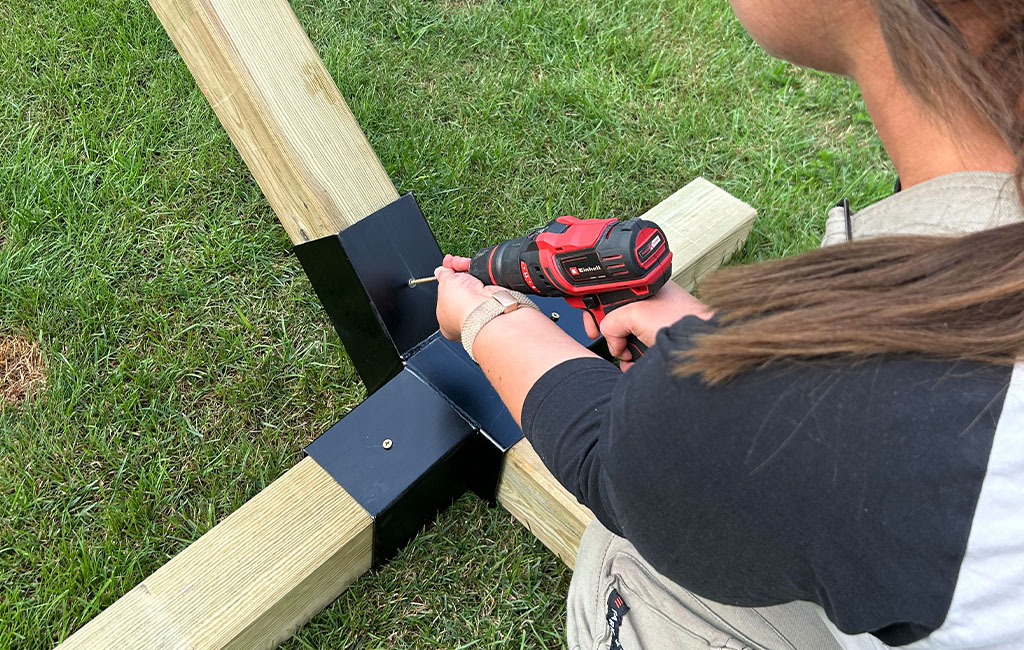 A woman uses an Einhell cordless drill to fasten a black metal bracket to a wooden cross frame.