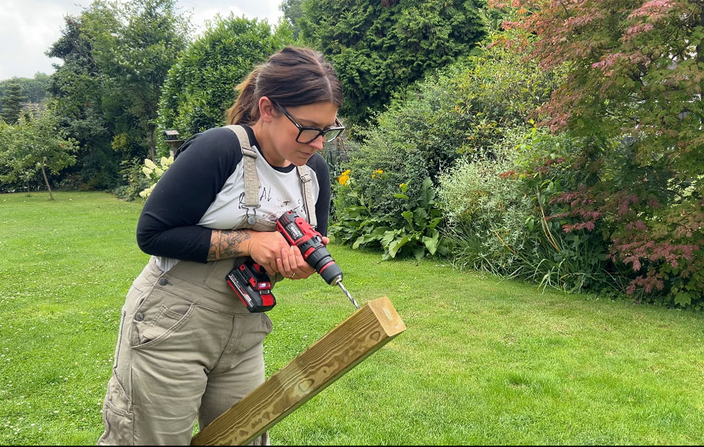 A woman drills a hole into the end of a wooden beam in the garden using an Einhell cordless drill.