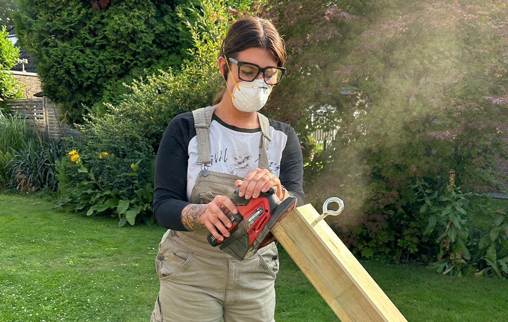 A woman sands a wooden beam with an Einhell cordless multi-sander; she wears safety goggles and a mask.