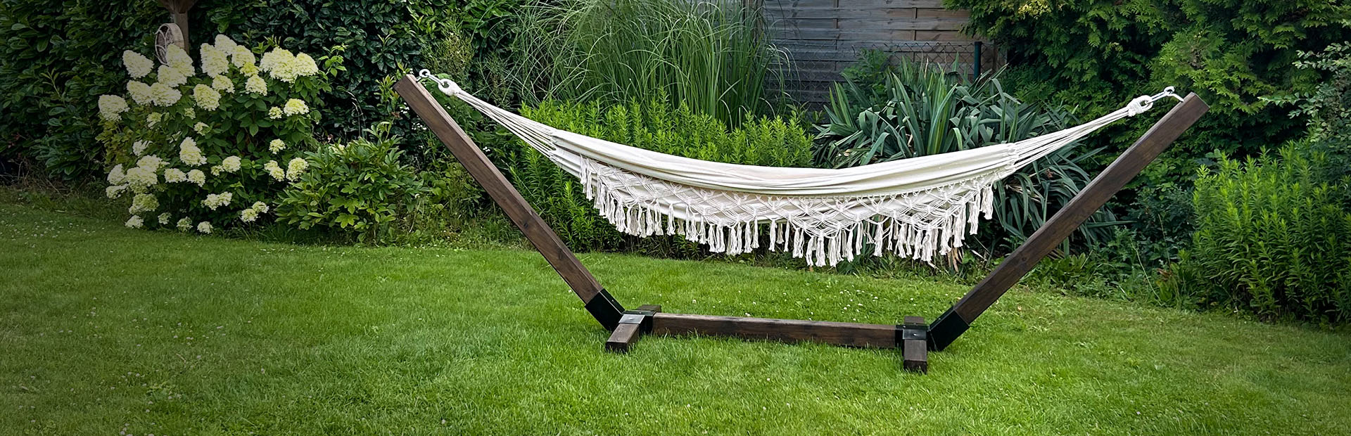 A cream-colored hammock with fringes hangs in a dark wooden frame on a lawn in a garden.