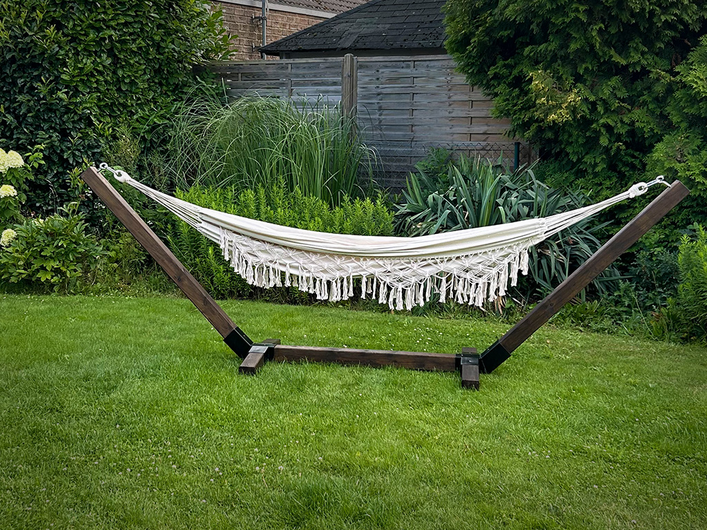 Eine cremefarbene Hängematte mit Fransen hängt in einem dunklen Holzgestell auf einer Rasenfläche im Garten.