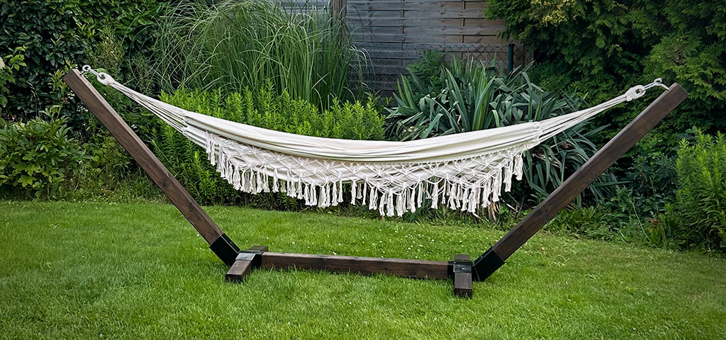 A cream-colored hammock with fringes hangs in a dark wooden frame on a lawn in a garden.