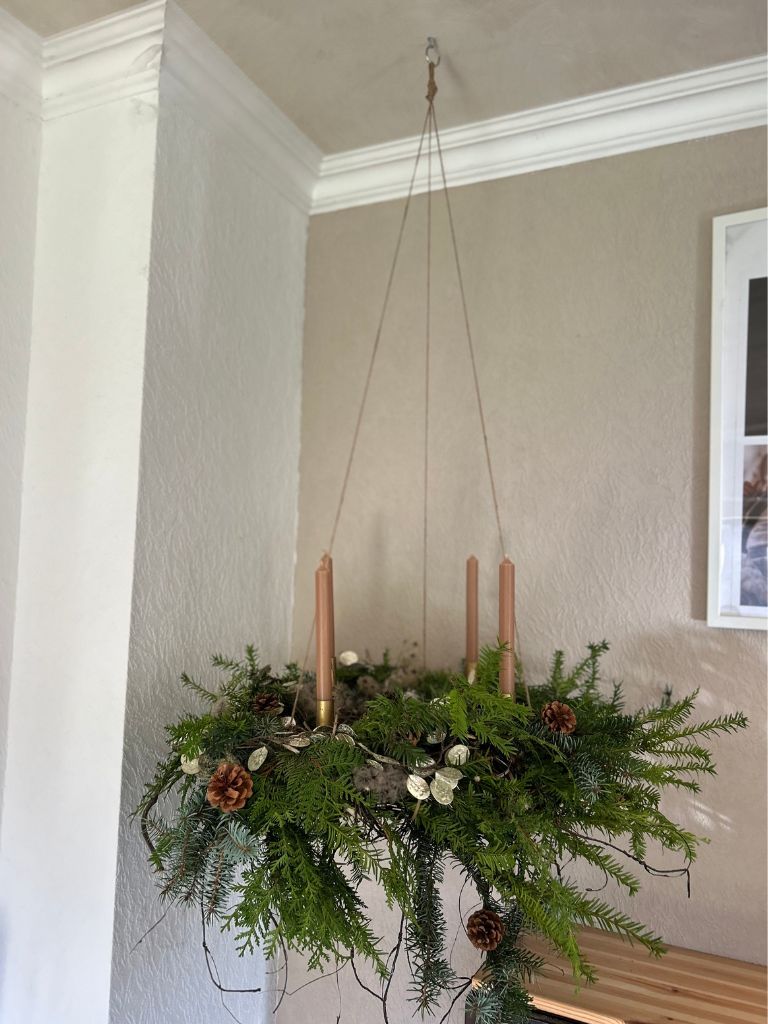 A hanging Advent wreath made of fir greenery with four brown taper candles, decorated with cones and dried plants.