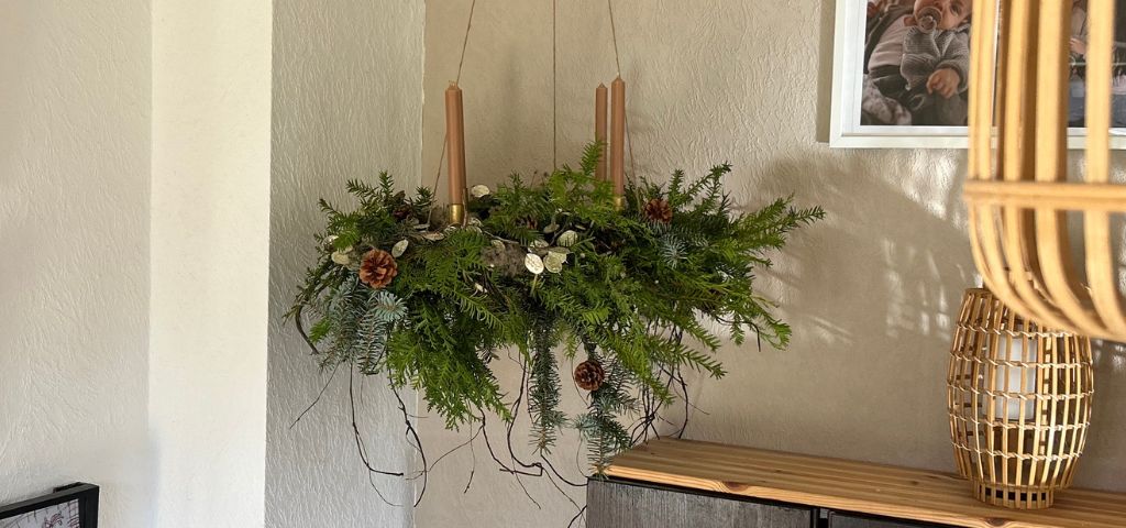 A hanging Advent wreath made of fir greenery with four brown taper candles, decorated with cones and dried plants.