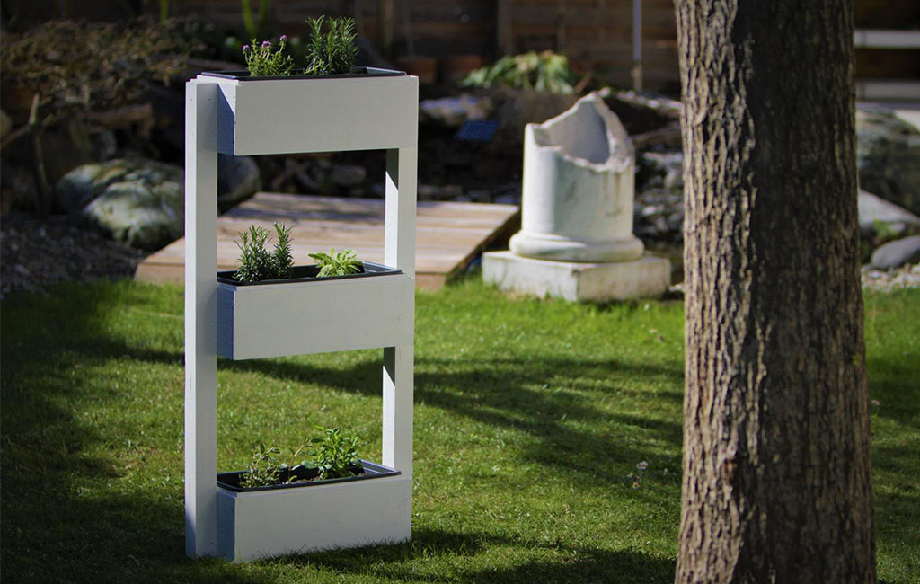 A white, rectangular herb garden with three stacked planting boxes in a garden. In the background, a tree, a broken stone column, and a wooden walkway.