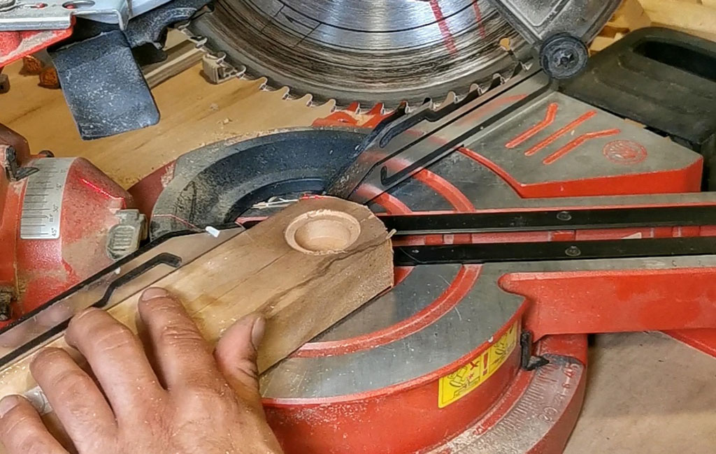 A hand positions a wooden piece with a round recess under a miter saw for further processing.