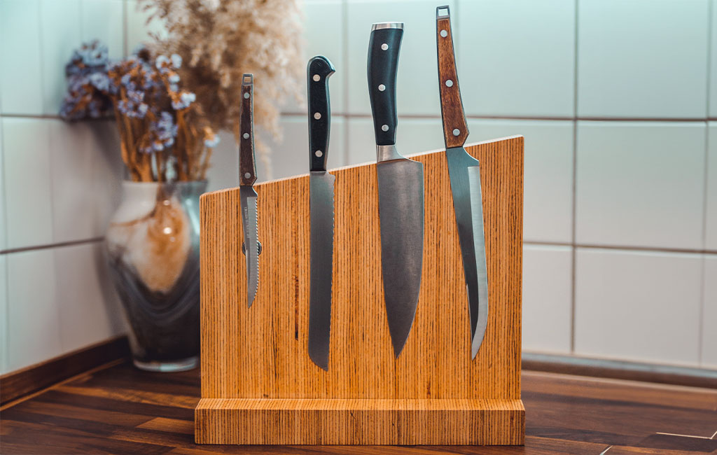 A wooden knife block with four different kitchen knives stands on a countertop in front of a tiled white wall.