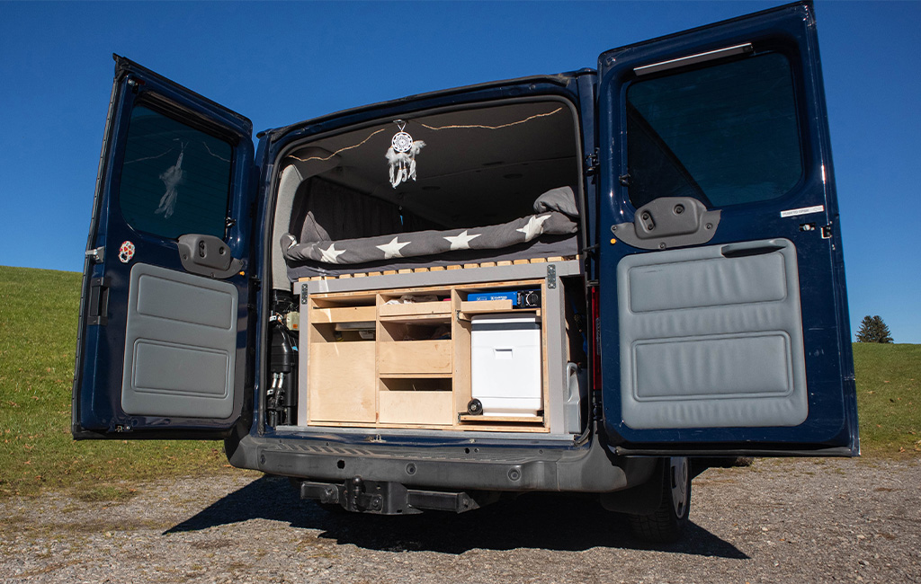 View into the open rear of a van with built-in wooden module, mattress, and storage space.