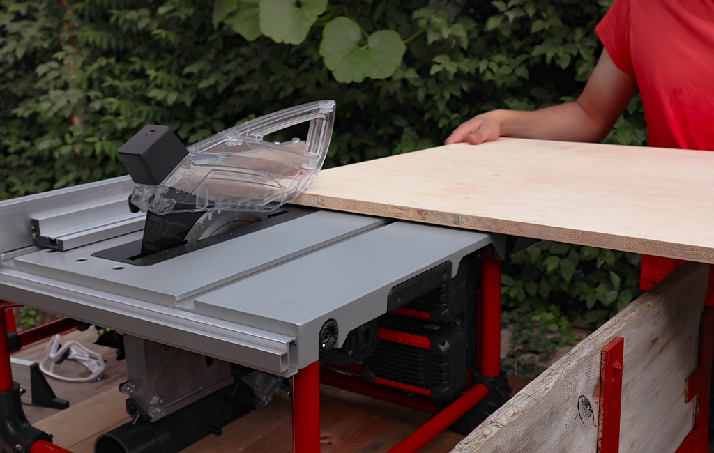 Person pushing a wooden board over a table saw blade with transparent safety guard.