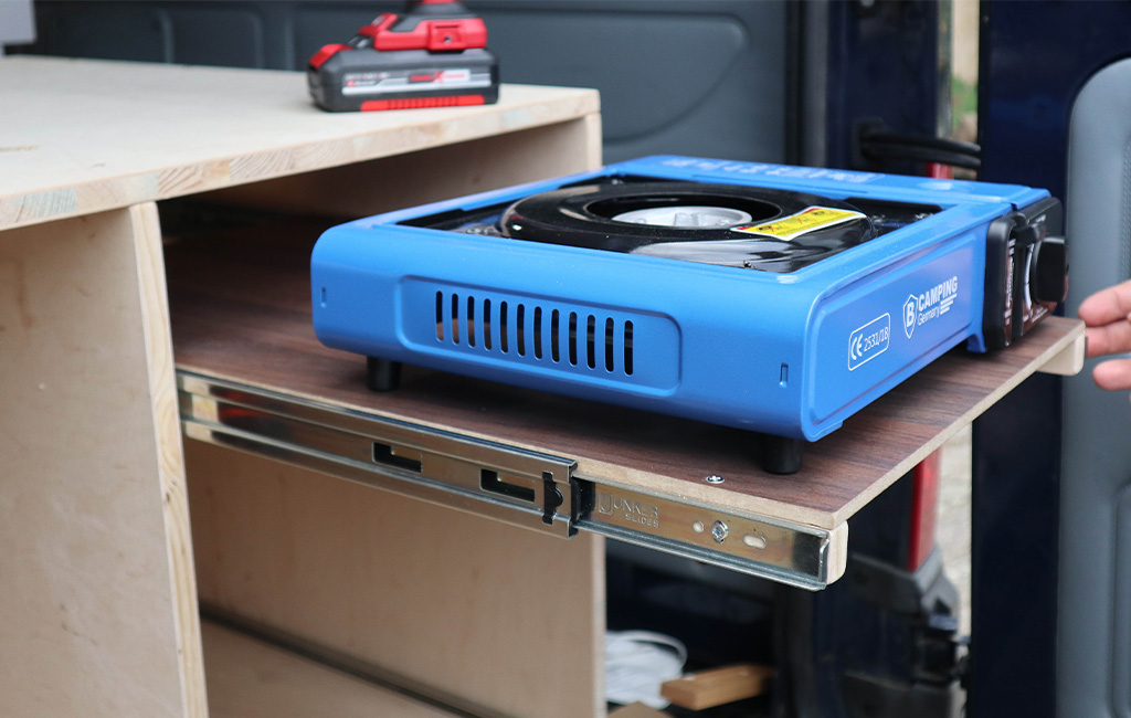 Blue camping stove on a pull-out wooden board with metal rail inside a camper.