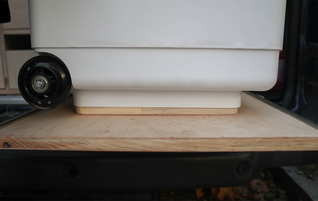 White canister with wheels fits exactly onto a wooden base in the rear of a van.