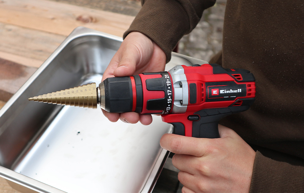 Einhell cordless drill with step drill bit held above a stainless steel sink.