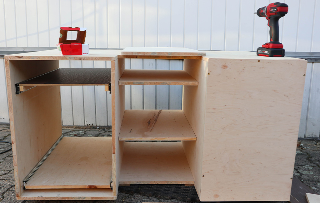 Partially assembled wooden module with shelves and drawer slide, cordless drill placed on top.