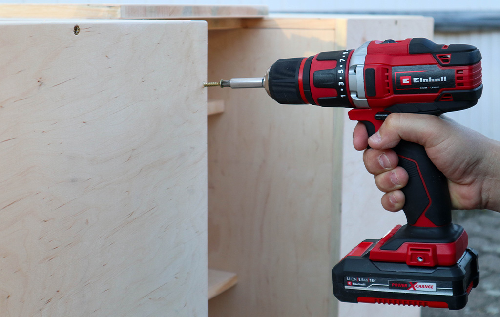 Einhell cordless drill driving a screw into the side panel of a wooden module.