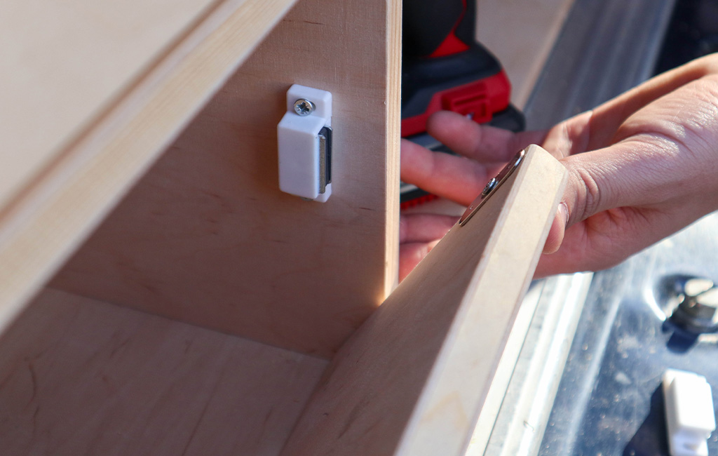 Magnetic catch being mounted to a wooden door, with a cordless drill visible in the background.