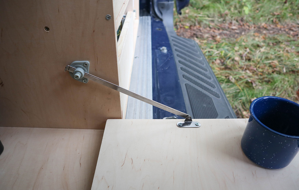 Folding mechanism with metal stay supports an opened wooden surface inside the van.