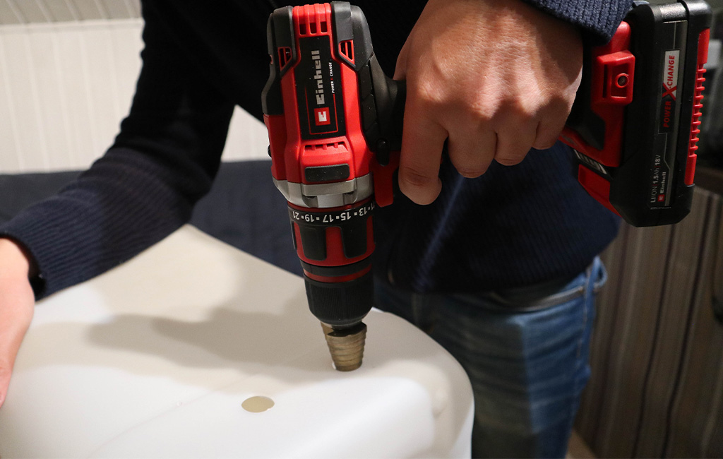 Einhell cordless drill with step drill bit making a hole in a white plastic canister.