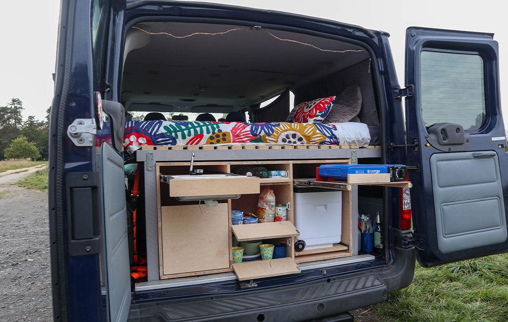 Open van with extended camping kitchen showing gas stove, sink, and food storage compartments.