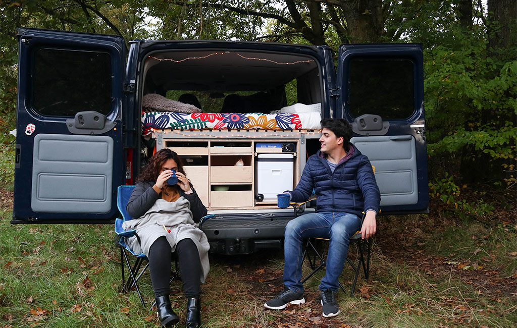 Two people relaxing with drinks in front of an open van, with a colorful bed and camping box in the background.