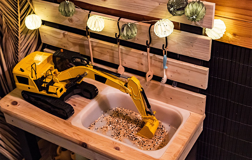A yellow toy excavator stands on the wooden top of the mud kitchen, digging in a sand-filled sink.