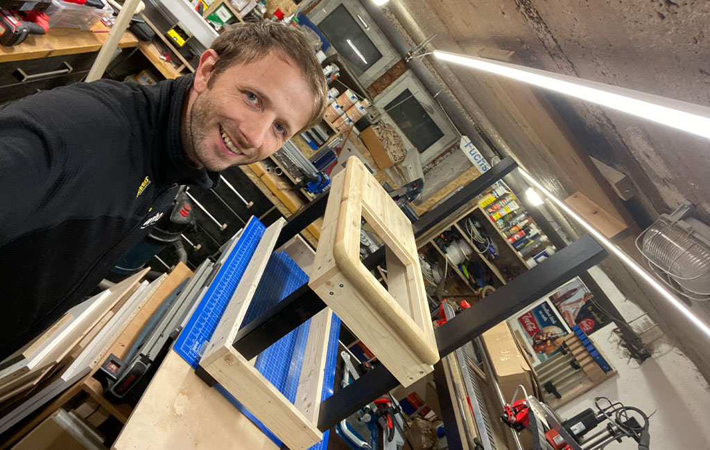 A man smiles at the camera while working on the wooden structure of the mud kitchen.