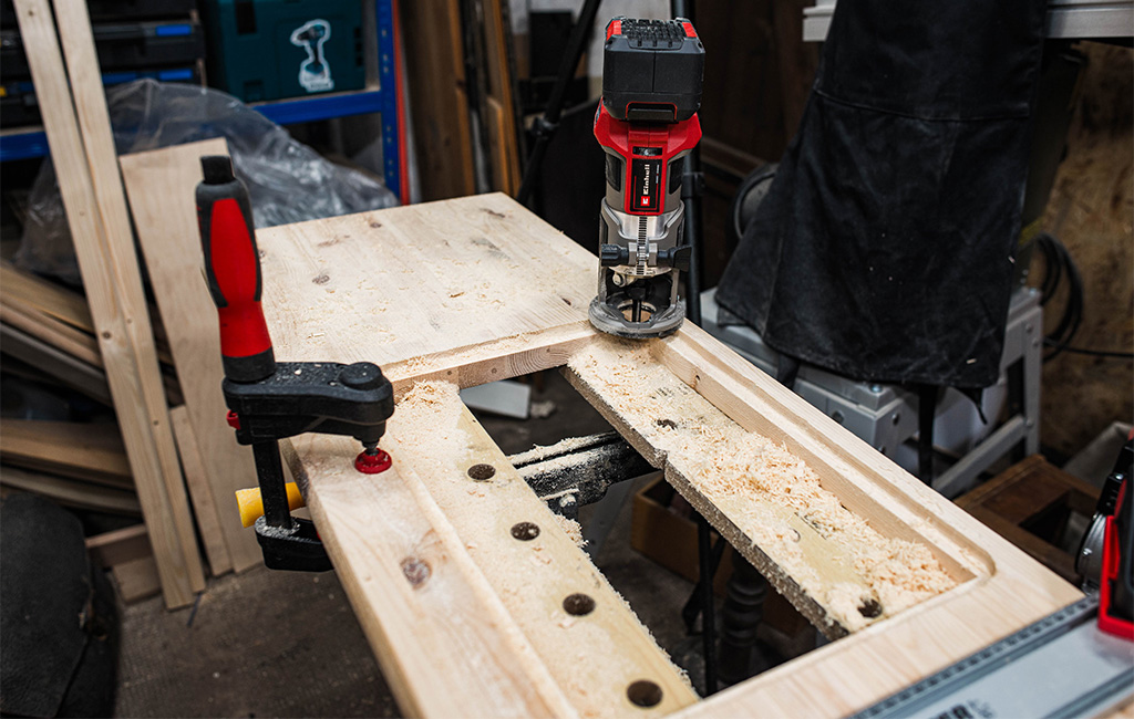 A cordless edge router is in use, routing a recess into a wooden board of the mud kitchen.
